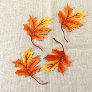 Op de afbeelding: Vier geborduurde esdoornblad patches in tinten oranje en geel. Elk blad heeft gedetailleerde stiksels en een bruine steel. De bladeren zijn gerangschikt op een neutraal gekleurde stof, perfect voor herfstthema knutselen.