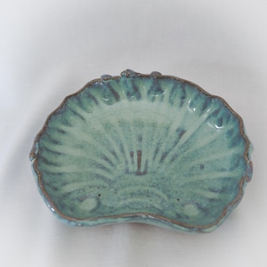 Puede incluir: Un pequeño plato de cerámica formado a mano con forma de concha de vieira. La superficie presenta un esmalte brillante y moteado en tonos verde espuma de mar y turquesa, con bordes marrón oscuro que resaltan el borde festoneado.