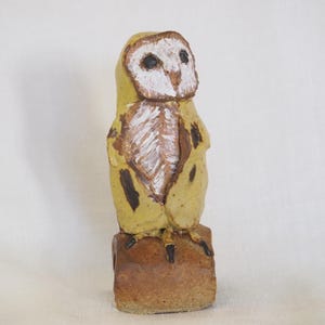 May include: A hand-sculpted ceramic owl figurine featuring a yellow body with brown spots, a white heart-shaped facial disc, and dark eyes. The owl stands on a rectangular brown base, showcasing a textured, rustic finish with visible brushstrokes.