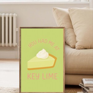 Könnte beinhalten: Gerahmter Druck mit hellgrünem Hintergrund, der ein Stück Key-Lime-Pie mit Schlagsahne zeigt. Der Text "YOU HAD ME AT" steht über dem Kuchen und "KEY LIME" darunter, alles in Rosa.