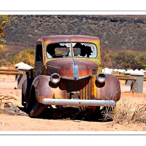 May include: An antique, rusty pickup truck with broken windshield glass. The truck is brown and orange, with two round headlights and a silver bumper. It is parked on a sandy surface with desert vegetation in the background.