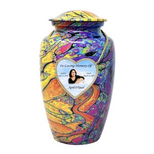 Pu&ograve; includere: Un'urna crematoria colorata con una targa commemorativa a forma di cuore. La targa contiene una foto di una donna e il testo "In Loving Memory Of", "Sunrise July 15, 1970", "Sunset March 25, 2023", e "April O'kneel".
