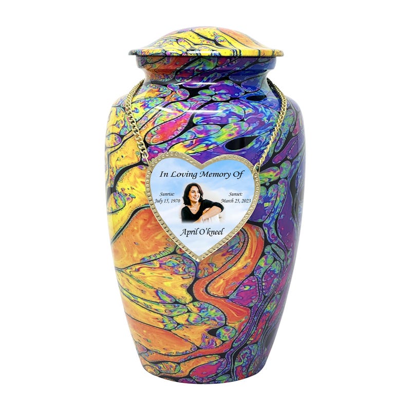 Pu&ograve; includere: Un'urna crematoria colorata con una targa commemorativa a forma di cuore. La targa contiene una foto di una donna e il testo "In Loving Memory Of", "Sunrise July 15, 1970", "Sunset March 25, 2023", e "April O'kneel".