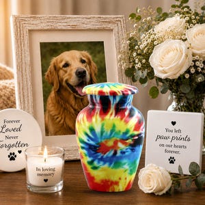 K&ouml;nnte beinhalten: Eine Urne aus Metall mit Tie-Dye-Muster und passendem Deckel, neben einem wei&szlig;en Kerzenhalter mit der Aufschrift In loving memory, einer runden Plakette mit der Aufschrift Forever Loved Never Forgotten und einer quadratischen Plakette.