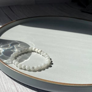 May include: A white moonstone beaded bracelet on a light grey ceramic plate. The bracelet is made of small, round beads and has a simple design.