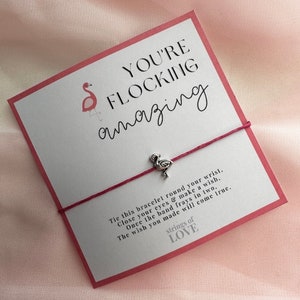 May include: A pink and white card with the text "You're flocking amazing" and a pink flamingo illustration. A red string bracelet with a silver flamingo charm is attached to the card. The card reads "Tie this bracelet round your wrist. Close your eyes & make a wish. Once the band frays in two, The wish you made will come true. strings of LOVE"