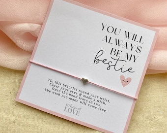 Peut inclure: Une carte rose avec le texte "You will always be my bestie" et un charm en forme de cœur argenté attaché à un bracelet en corde. La carte comprend également des instructions pour faire un vœu et le texte "strings of love".