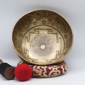 May include: A golden singing bowl with intricate geometric patterns, resting on a patterned cushion. A wooden mallet with a black handle and a red felt striker are placed nearby. The bowl's design includes a central mandala.