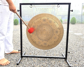 May include: A large circular brass gong suspended from a black metal frame. The gong features intricate etched patterns, including a central square mandala design surrounded by circular motifs and Sanskrit text. A wooden mallet with a red felt head is used.