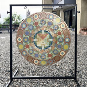 May include: A large circular metal gong suspended from a black rectangular metal stand. The gong features a central square mandala design surrounded by concentric rings of small, colorful, intricately patterned circular motifs in shades of blue, red, yellow, and green.
