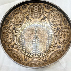 May include: A gold and black singing bowl with intricate designs, including a flower of life pattern and two footprints. The bowl is made of metal and has a smooth, polished surface.