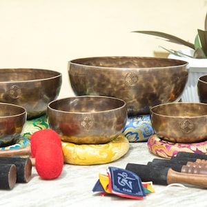 Puede incluir: Conjunto de seis cuencos cantores de aspecto antiguo con acabado martillado. Los cuencos están dispuestos en orden de tamaño, con el cuenco más grande en la parte superior y el más pequeño en la parte inferior. Cada cuenco tiene un pequeño diseño circular en el interior. Los cuencos están sentados sobre cojines coloridos. Hay tres mazos de madera con puntas de goma negra.