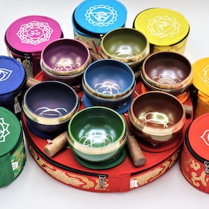 May include: A set of seven singing bowls, each with a different colour and design. Each bowl is in a separate pouch with a zipper closure. The pouches are decorated with the seven chakra symbols. The bowls are arranged in a circle on a red cushion with a gold trim. The bowls come with two wooden mallets.