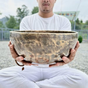 Pode incluir: Uma taça tibetana de bronze martelado, grande e redonda, com uma pátina escura e manchada. A superfície exterior apresenta um símbolo circular gravado e centrado, composto por quatro quadrantes simétricos dentro de um rebordo fino e escuro.