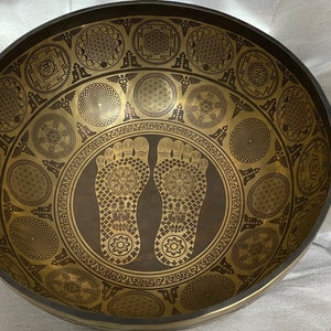 May include: A large, round, brown singing bowl with a gold design. The bowl features a pattern of intricate circles and symbols, including two large footprints in the centre.