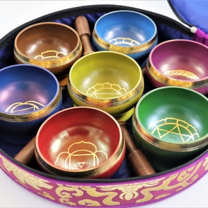 Op de afbeelding: Set van zeven zangkommen in een paarse draagtas. De kommen zijn in een cirkel gerangschikt en zijn elk van een andere kleur: paars, bruin, geel, roze, groen, rood en blauw. Elke kom heeft een gouden ontwerp aan de binnenkant.