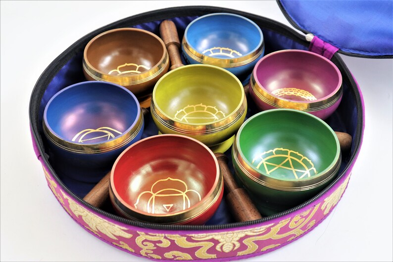 Op de afbeelding: Set van zeven zangkommen in een paarse draagtas. De kommen zijn in een cirkel gerangschikt en zijn elk van een andere kleur: paars, bruin, geel, roze, groen, rood en blauw. Elke kom heeft een gouden ontwerp aan de binnenkant.