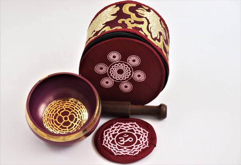 Op de afbeelding: Een bordeauxrode zangkomset met een gouden mandala-ontwerp, een houten slaagstok en een bordeauxrood kussen met een wit Om-symbool.