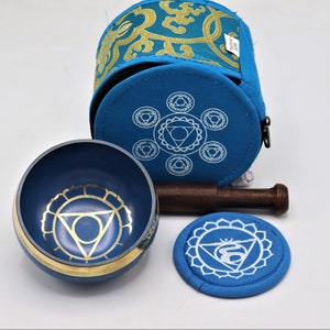 Op de afbeelding: Een blauwe zangkomset met een houten slaagstok, een blauw kussen en een blauwe draagtas met een ritssluiting. De kom en het kussen hebben een gouden en wit ontwerp met een chakra-symbool.