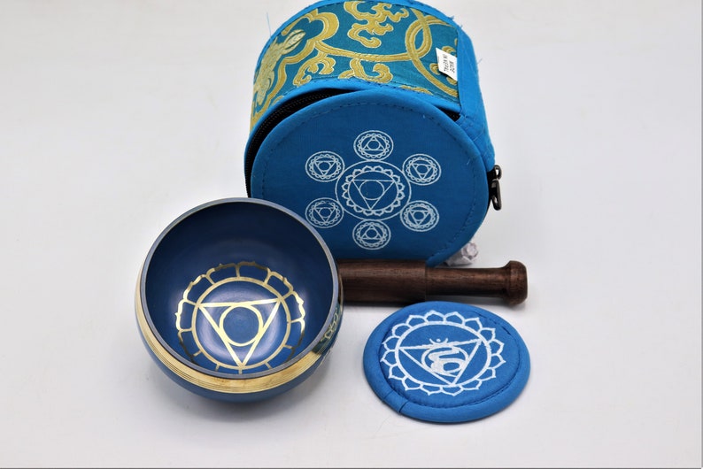 Op de afbeelding: Een blauwe zangkomset met een houten slaagstok, een blauw kussen en een blauwe draagtas met een ritssluiting. De kom en het kussen hebben een gouden en wit ontwerp met een chakra-symbool.