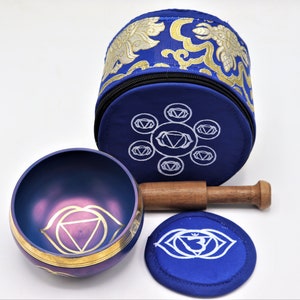 Op de afbeelding: Een blauwe zangkom met een gouden rand en een paars derde oog chakra symbool. De kom wordt geleverd met een houten slaagstok en een blauw kussen met het derde oog chakra symbool.