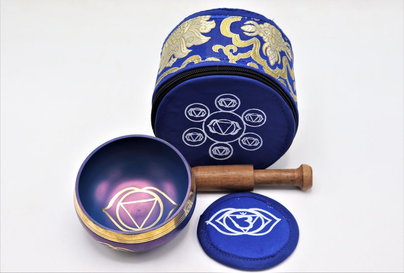 Op de afbeelding: Een blauwe zangkom met een gouden rand en een paars derde oog chakra symbool. De kom wordt geleverd met een houten slaagstok en een blauw kussen met het derde oog chakra symbool.
