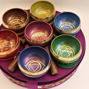 Op de afbeelding: Set van zeven zangkommen in verschillende kleuren met gouden ontwerpen, gerangschikt in een cirkel op een paarse draagtas.