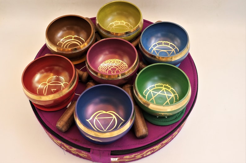 Op de afbeelding: Set van zeven zangkommen in verschillende kleuren met gouden ontwerpen, gerangschikt in een cirkel op een paarse draagtas.
