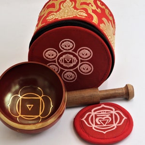 Op de afbeelding: Een set van zeven chakra-zingende kommen met een houten slaagstok en een rode fluwelen draagtas. De kommen zijn versierd met gouden en roze chakra-symbolen.