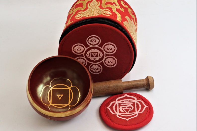 Op de afbeelding: Een set van zeven chakra-zingende kommen met een houten slaagstok en een rode fluwelen draagtas. De kommen zijn versierd met gouden en roze chakra-symbolen.