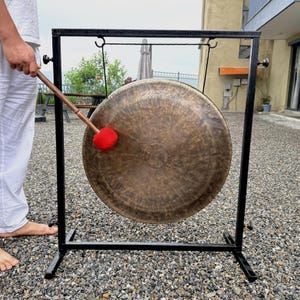 May include: A large, circular, bronze-colored metal gong suspended from a black rectangular metal frame. The gong has a textured, hammered surface and is being struck by a wooden mallet with a bright red, spherical felt head.