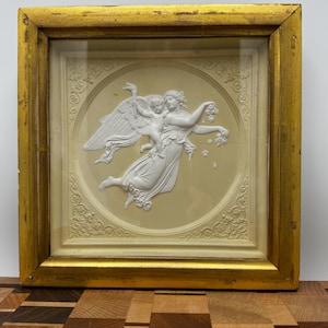 May include: A framed white plaster relief sculpture of a winged woman holding a cherub. The sculpture is set against a cream background with a gold frame.