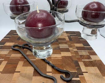 Puede incluir: Un candelabro de hierro forjado con cuatro cuencos de vidrio, cada uno con una vela grande y redonda de color burdeos. Dos ganchos de hierro forjado están sobre una tabla de cortar de madera con un patrón geométrico. Un acento decorativo para el hogar.