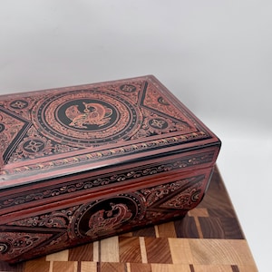 Pode incluir: Uma caixa decorativa retangular vermelha e preta com padrões intrincados e um desenho de fénix na tampa e na frente. A caixa é feita de madeira e tem um acabamento brilhante. A caixa está sobre uma tábua de cortar de madeira.