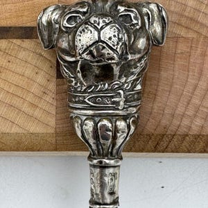 May include: A silver-toned metal cane handle shaped like a pug dog head. The dog features a detailed face, floppy ears, and a collar with a buckle. Below the head is a fluted, bell-shaped transition piece that connects to a dark, cylindrical shaft.