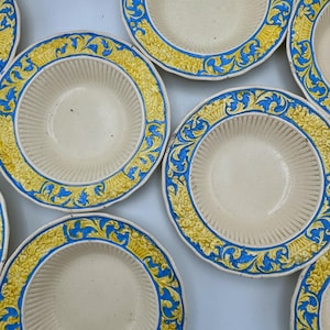 Puede incluir: Una colección de cuencos de cerámica redondos con un centro de color crema y acanaladuras verticales. Los bordes anchos y planos están decorados con un patrón en relieve ornamentado en amarillo vibrante y azul, con volutas detalladas.