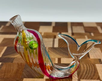 May include: A glass mermaid figurine with a colorful striped tail and a green glass bead for an eye. The figurine is sitting on a wooden surface.