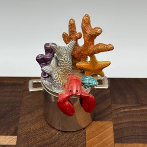 Puede incluir: Una olla de plata con una langosta roja, un pez plateado y coral naranja, morado y amarillo en la parte superior.