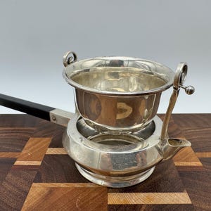 Op de afbeelding: Een theezeef van verzilverd metaal met een gepolijste afwerking, voorzien van een diepe kom, een kleine tuit en een lange, rechte zwarte handgreep. De zeef is ontworpen met twee decoratieve metalen lussen aan de zijkanten en rust op een ronde voet.