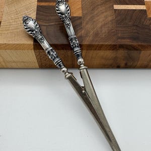 May include: A pair of silver-toned metal sugar tongs featuring ornate, embossed handles with intricate scrollwork and floral detailing. The tongs have long, slender, pointed tips that meet at a single point, showcasing a polished metallic finish.