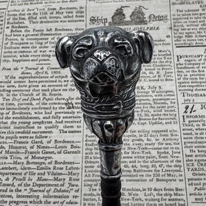 Puede incluir: Un bastón de paseo plateado con un mango de cabeza de perro pug detallado. El bastón está hecho de madera y tiene un acabado negro. El mango está decorado con una hebilla de cinturón de plata. El bastón está apoyado en un periódico con texto de la década de 1800.