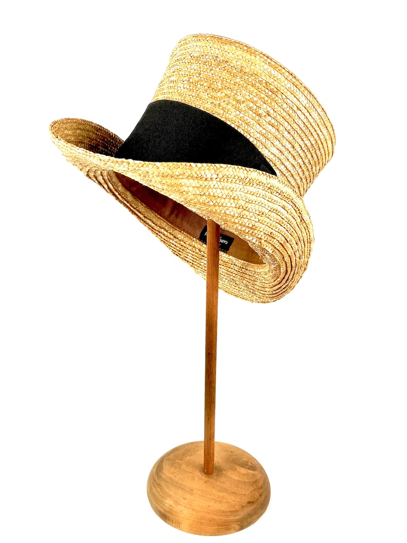 May include: A straw hat with a black band, displayed on a wooden stand. The hat has a tall, cylindrical crown and a wide brim.
