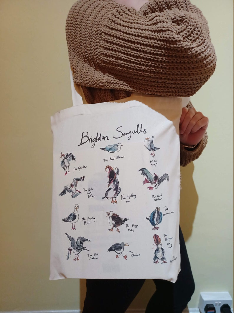 May include: A white canvas tote bag featuring a black and white illustration of seagulls with the text "Brighton Seagulls". The illustration includes various types of seagulls with names such as "The Gouder", "The Road Blacher", "The Gobbay", "The Flotty Baby", and "The Fish Snatcher".