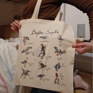 May include: A white canvas tote bag with a brown handle. The bag features a colourful illustration of seagulls with names and descriptions. The text "Brighton Seagulls" is printed in brown at the top of the bag.
