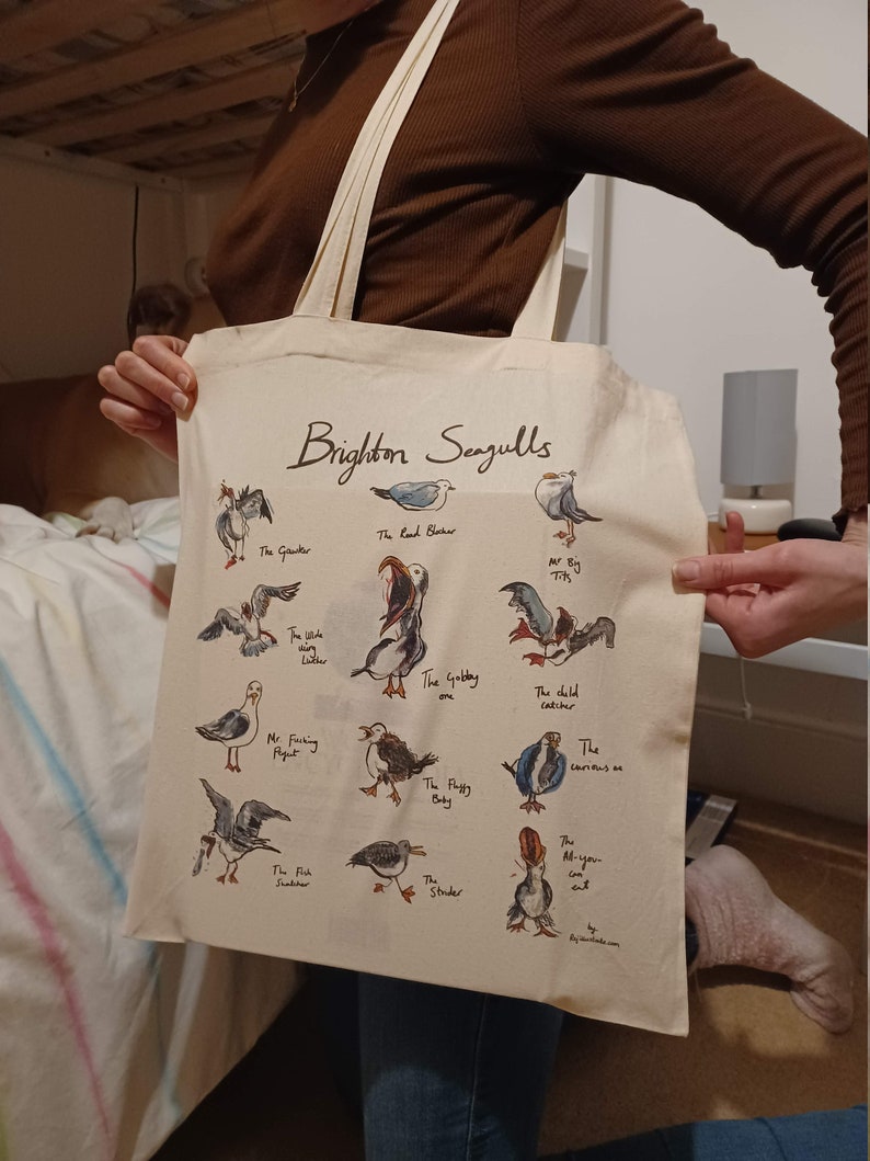 May include: A white canvas tote bag with a brown handle. The bag features a colourful illustration of seagulls with names and descriptions. The text "Brighton Seagulls" is printed in brown at the top of the bag.