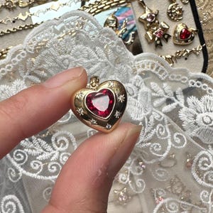 Peut inclure: Un pendentif en forme de cœur doré avec un centre en pierre rouge et de petits accents en forme d'étoile. Le pendentif est présenté sur un fond d'autres bijoux et de dentelle blanche.