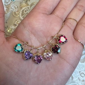 May include: A gold necklace with seven heart-shaped pendants in teal, purple, pink, and red. Each pendant has a gemstone. The necklace is held in a hand, with a fine chain. The background is a lace fabric.