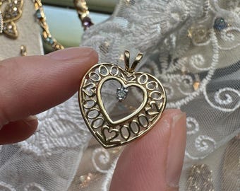 Könnte beinhalten: Goldfarbener herzförmiger Anhänger mit einem kleinen Diamanten in der Mitte. Der Anhänger hat die Zahlen "2000" und "2007" um das Herz herum eingraviert. Der Anhänger wird von den Fingern einer Person gehalten.