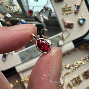 May include: A gold pendant with an oval, deep red gemstone is held between fingers. Other gold jewellery, including necklaces and charms, are visible in a jewellery box. The gemstone is the focal point.