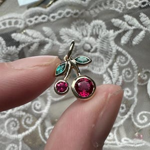 May include: A gold cherry pendant featuring two red gemstones as cherries and two green gemstones as leaves. The pendant is held between two fingers.